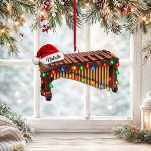 Könnte beinhalten: Ein hängendes Xylophon-Ornament mit einer roten Weihnachtsmannmütze, auf der der Name Natalie in schwarzer Schrift steht. Das Instrument hat braune Holzklangplatten, goldfarbene Resonanzrohre und eine bunte Lichterkette.