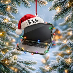 May include: A silver laptop ornament featuring a red and white Santa hat with the name Natalie printed in black script. The laptop is wrapped in a string of multicoloured Christmas lights and hangs from a thin red ribbon against a snowy background.
