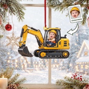 May include: A yellow and black excavator ornament featuring a baby wearing a yellow hard hat in the driver's seat. The ornament hangs from a red ribbon and includes a small inset box showing a baby in a hard hat with the text UPLOAD PHOTO.