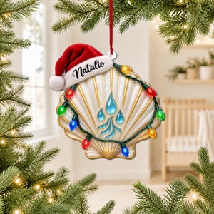 Puede incluir: Un adorno navideño con forma de concha de vieira que lleva un gorro de Papá Noel rojo con el nombre Natalie impreso en el borde blanco. La concha está decorada con una cadena de luces navideñas de colores y un diseño central de gotas de agua azules.