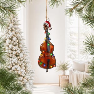 May include: A brown wooden cello ornament hanging from a string, decorated with a string of colorful Christmas lights. The cello features a red and white Santa hat on its scroll with the name Natalie printed on the white trim of the hat.