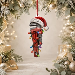 May include: A red pipe wrench hanging ornament featuring a Santa hat with the name Natalie printed on the white trim. The tool is wrapped in a string of multicolored Christmas lights and suspended by a thin red ribbon for holiday decoration.
