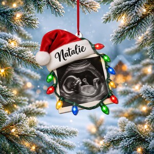 May include: A square-shaped Christmas ornament featuring a black and white ultrasound image of a fetus. The ornament is topped with a red and white Santa hat with the name Natalie printed on the brim and is wrapped in a string of colorful lights.