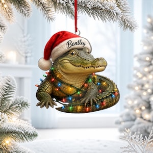 Op de afbeelding: Een hangende kerstversiering met een groene alligator die een rood-witte kerstmuts draagt met de naam Bentley op de rand. De alligator is omwikkeld met een snoer van kleurrijke, gloeiende kerstverlichting en hangt aan een rood lint.