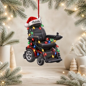 May include: A hanging Christmas ornament shaped like a black power wheelchair decorated with colorful string lights. The wheelchair features a red Santa hat on the headrest with the name Natalie printed in black script and hangs from a red ribbon.