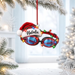 Könnte beinhalten: Ein hängender Weihnachtsschmuck in Form einer Schwimmbrille mit blauen Gläsern, umwickelt mit einer Lichterkette aus bunten Weihnachtslichtern und gekrönt von einer roten Weihnachtsmannmütze mit dem Namen Natalie in schwarzer Schrift.