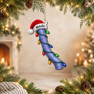 Può includere: Un ornamento curvo, testurizzato e viola chiaro a forma di tubo da piscina, sormontato da un cappello da Babbo Natale rosso e bianco con il nome Natalie in caratteri neri. L'ornamento è avvolto da una catena di luci natalizie colorate.