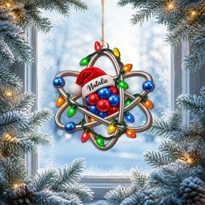 Op de afbeelding: Een hangend ornament in de vorm van een atoom met zilverkleurige metalen banen, een centrale kern van rode en blauwe bollen en een rode kerstmuts met de naam Natalie. Het geheel is omwikkeld met een snoer van gekleurde kerstverlichting.