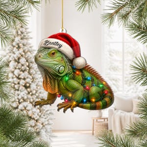 Puede incluir: Un adorno navideño colgante que muestra una iguana verde con un gorro de Papá Noel rojo y blanco con el nombre Bentley impreso en el borde. La iguana está envuelta en luces de colores brillantes y suspendida por un cordón dorado.