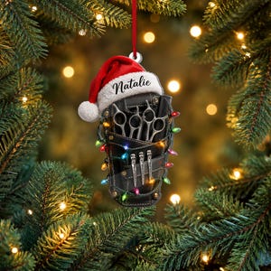 May include: A black leather-look barber tool belt ornament featuring a red Santa hat with the name Natalie printed on the white trim. The belt holds silver scissors, a comb, and hair clips, all wrapped in colourful miniature Christmas lights.