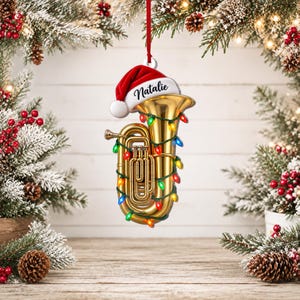 Peut inclure: Une décoration de Noël en forme de tuba doré portant un bonnet de Père Noël rouge avec le prénom Natalie inscrit en lettres noires. L'instrument en laiton est entouré d'une guirlande lumineuse multicolore et suspendu par un ruban rouge.