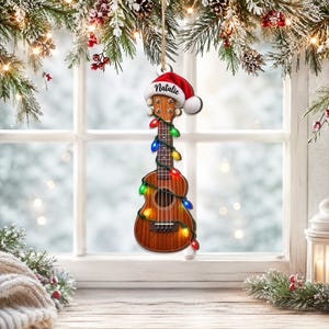 Könnte beinhalten: Ein hölzernes Ukulele-Ornament mit einer rot-weißen Weihnachtsmannmütze auf dem Kopf, auf deren Rand der Name Natalie steht. Das Instrument ist mit einer Lichterkette aus bunten, leuchtenden Weihnachtslichtern umwickelt und hängt an einer Schnur.