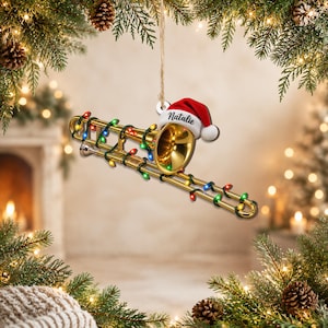 Puede incluir: Un adorno de trombón dorado colgado de una cuerda, decorado con una cadena de luces navideñas multicolores. Un gorro de Papá Noel rojo y blanco se asienta sobre la campana del instrumento, con el nombre Natalie impreso en letra negra.