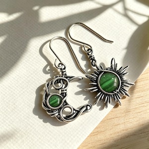 May include: A pair of silver-toned dangle earrings featuring a crescent moon and a sun design. Each earring is crafted with intricate wire-wrapped metalwork and contains a round, smooth, green stone set in the center of the celestial shapes.