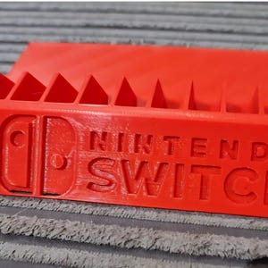 May include: A bright red plastic storage rack for game cartridges featuring the Nintendo Switch logo embossed on the front. The rectangular stand includes a series of vertical slots along the top edge designed to hold multiple game cases securely.