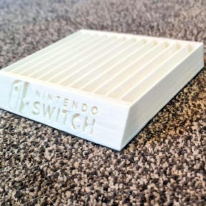 May include: A white, 3D-printed plastic storage rack for Nintendo Switch game cartridges. The rectangular organiser features multiple parallel slots for holding game cases and displays the Nintendo Switch logo embossed on the front slanted edge.