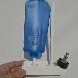 Pode incluir: Um recipiente de água azul Softflask de 500 ml montado num suporte de secagem de plástico branco. O suporte possui um poste de apoio vertical e uma bandeja base. Uma tampa preta e cinzenta está posicionada no lado direito da base.