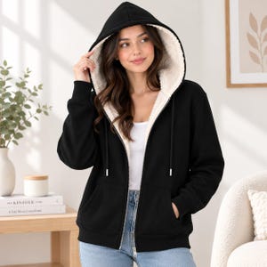 May include: A black zip-up hoodie featuring a white sherpa fleece lining, a hood with matching drawstrings, and two front pockets. The garment is worn over a white top and blue jeans, showcasing a full-length front zipper and ribbed cuffs.
