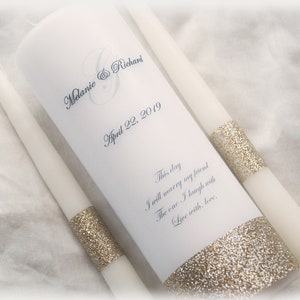 Puede incluir: Dos velas de unidad de boda blancas con ribete de purpurina dorada. La vela más grande tiene los nombres "Melanie & Richard" y la fecha "April 22, 2019" impresos en ella. El texto "This day I will marry my friend The one I laugh with Live with, love" también está impreso en la vela más grande.