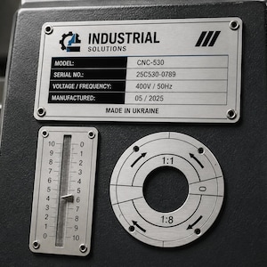 Peut inclure: Un panneau de machine industriel gris foncé avec une plaque d'identification argentée portant le texte INDUSTRIAL SOLUTIONS, modèle CNC-530, série 25C530-0789, 400V/50Hz et 05/2025. En dessous se trouvent une échelle linéaire verticale de 0 à 10 et un cadran.