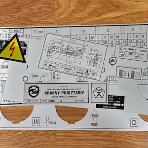 May include: A collection of industrial machine data plates and control panel overlays featuring technical diagrams, electrical warning symbols, and text including "Krasny Proletariy" and "Made in USSR." The metallic labels display charts, numerical data, and circular cutouts for machinery controls.