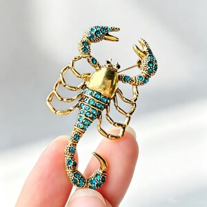 Op de afbeelding: Een goudkleurige schorpioenbroche met een gladde, gepolijste kop en een getextureerd lichaam versierd met talloze kleine, felblauwe strass-steentjes. De schorpioen heeft twee opgeheven scharen en een gekrulde staart met een speld aan de achterzijde.