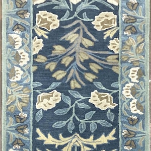 May include: A rectangular wool runner rug featuring a deep navy blue background with a symmetrical floral pattern in shades of light blue, sage green, and cream. The design includes a central botanical motif surrounded by a decorative floral border.