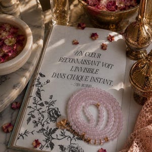 Peut inclure: Un collier de perles rondes rose pâle avec des accents dorés repose sur un livre ouvert orné d'illustrations florales noires et du texte : UN COEUR RECONNAISSANT VOIT L'INVISIBLE DANS CHAQUE INSTANT. - IBN RUSHD, entouré de pétales éparpillés.