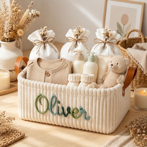 May include: A rectangular cream-colored corduroy storage basket with the name Oliver embroidered in green yarn on the front. The basket contains a beige long-sleeve baby shirt, three white drawstring pouches, two bottles, baby booties, and a teddy bear.