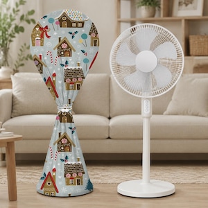 Peut inclure: Un ventilateur sur pied blanc à côté d'une housse en tissu avec un fond bleu clair orné d'un motif répétitif de maisons en pain d'épices marron, de sucres d'orge rouges et blancs, de sucettes colorées, de houx vert et de flocons blancs.