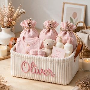 Op de afbeelding: Een witte mand van corduroy met de naam Oliver geborduurd in roze schrift. Binnenin zitten drie roze mousseline zakjes met trekkoord, een roze shirt met lange mouwen en knoopjes, een kleine beige teddybeer, twee witte pompflessen en een kaartje met Happy 1st Birthday.