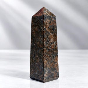 May include: A polished stone obelisk featuring a mottled texture with deep brown, black, and copper-coloured mineral inclusions. The tower has a hexagonal base and a pointed pyramidal top, showcasing a natural crystalline pattern throughout its surface.