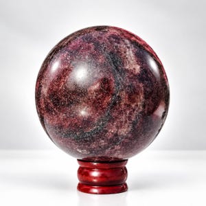 Puede incluir: Una piedra pulida en forma de esfera con un patrón jaspeado en color burdeos profundo y morado oscuro, con vetas negras sutiles y una superficie brillante y reflectante. La esfera descansa sobre un pequeño soporte de madera marrón rojizo.