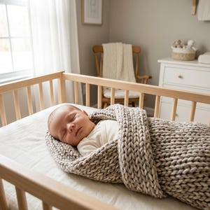 May include: A sleeping infant wrapped in a thick, chunky knit blanket in a neutral beige tone. The baby wears a simple cream-colored onesie and rests inside a light-colored wooden crib with vertical slats, lying on a plain white mattress.