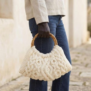 May include: A cream-colored cable knit handbag featuring a textured, chunky wool pattern and a rounded bamboo handle. The bag has a soft, slouchy structure with intricate braided cable details across the front surface, providing a tactile appearance.