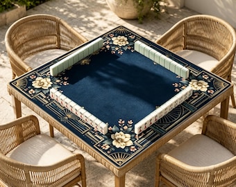 Könnte beinhalten: Ein quadratischer Mahjong-Tisch mit einer dunkelblauen Filzoberfläche, die an den Ecken cremefarbene Blumenmuster aufweist. Der Tisch ist von vier hellbraunen Stühlen aus geflochtenem Rattan mit cremeweißen Sitzkissen umgeben.
