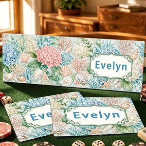 Può includere: Un cartello rettangolare e due sottobicchieri abbinati con una stampa ad acquerello di ortensie rosa e blu, conchiglie, corallo e perle. Ogni articolo mostra il nome Evelyn in testo blu all'interno di una cornice decorativa verde.