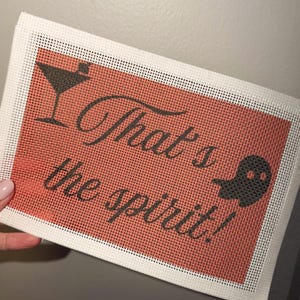 May include: A rectangular needlepoint canvas featuring a burnt orange background with a black martini glass icon on the top left and a black ghost icon on the right. The text "That's the spirit!" is printed in black cursive script in the center.