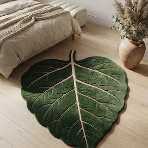 Peut inclure: Un tapis de sol vert foncé en forme de grande feuille avec une nervure centrale beige clair proéminente et des nervures plus petites. Le tapis présente une surface texturée et est posé sur un parquet clair. Le texte AARAV RUGS est visible.