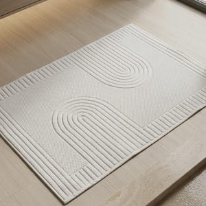 Può includere: Un tappetino da bagno rettangolare color bianco sporco con una superficie testurizzata caratterizzata da due motivi ad arco in rilievo specchiati. Il design include un bordo a linee verticali e una sezione centrale con una trama a griglia.