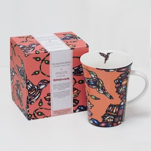 Peut inclure: Un mug en porcelaine et sa boîte carrée assortie, tous deux ornés d'un fond orange vif avec un motif coloré de colibris et de fleurs. Le mug présente un intérieur blanc avec un petit colibri près du bord et une anse blanche.