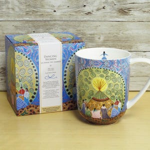 May include: A ceramic mug and its matching box featuring the Dancing Women design. The artwork displays a group of figures in colourful dresses circling a central yellow sun-like motif against a blue background with intricate swirling patterns.