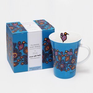 Peut inclure: Un mug en céramique bleue et sa boîte assortie, tous deux ornés d'un motif floral et d'oiseaux colorés. La boîte porte le texte FLOWERS AND BIRDS, OISEAUX ET FLEURS et Norval Morrisseau. Le mug a un intérieur blanc avec un petit motif d'oiseau.