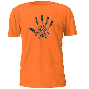 May include: A bright orange short-sleeved t-shirt featuring a dark green graphic on the chest. The graphic depicts a human hand silhouette filled with intricate nature-inspired illustrations, including feathers, flowers, and a pine tree.