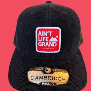 May include: Black corduroy baseball cap with a mesh back. The front features a red and white patch with the text "Ain't Life Grand Colorado". A metallic tag below reads "Cambridge" and "PB235".