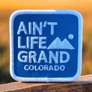 May include: A blue square patch with white stitching around the edges. The patch says "Ain't Life Grand Colorado" in white text with a white mountain peak graphic.