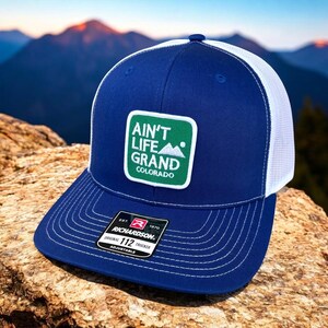 May include: A blue and white trucker hat with a green patch that says "Ain't Life Grand Colorado". The hat is made by Richardson 112.
