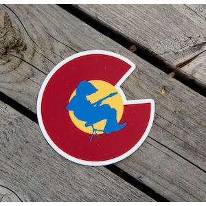 May include: A sticker featuring the Colorado state flag design. The sticker has a red background with a white border, a yellow circle, and a blue silhouette of a person playing a guitar. The sticker is placed on a wooden surface.