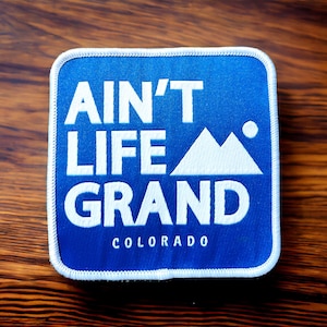 May include: A blue square patch with white stitching around the edges. The patch says "Ain't Life Grand" with a white mountain range graphic and the word "Colorado" below.