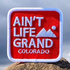 May include: Red square patch with white embroidered text that reads "Ain't Life Grand Colorado" with a white mountain silhouette in the center.